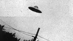Fotografía antigua en blanco y negro de un objeto discoidal en el cielo, junto a un poste y cables eléctricos.